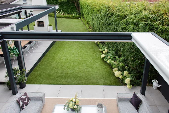 Jardín moderno con pérgola retráctil, césped, plantas y zona de estar al aire libre.