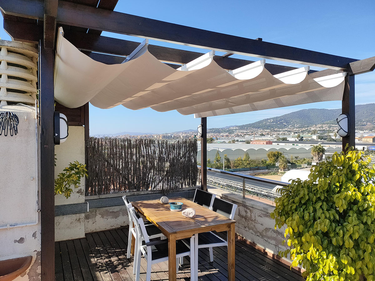 Terraza con mesa de madera, sillas y toldo, vista a montañas y cielo azul.