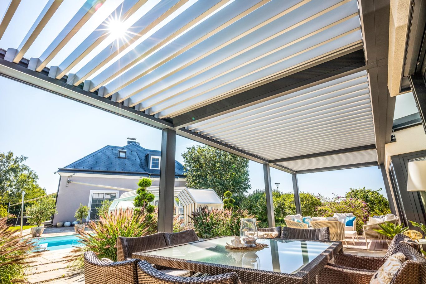 Terraza con pérgola, mesas y sillas, junto a una piscina y casa con techo a dos aguas.